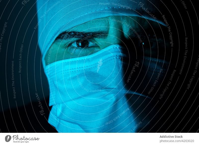 Male doctor in mask in dark room with blue neon light surgeon man medical male medicine protect illuminate sterile hospital treat clinic serious frown focus