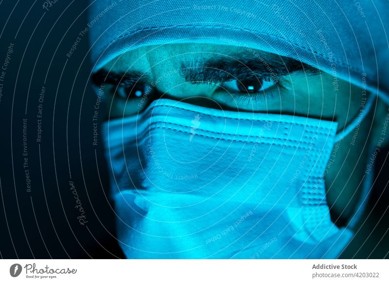 Male doctor in mask in dark room with blue neon light surgeon man medical male medicine protect illuminate sterile hospital treat clinic serious frown focus