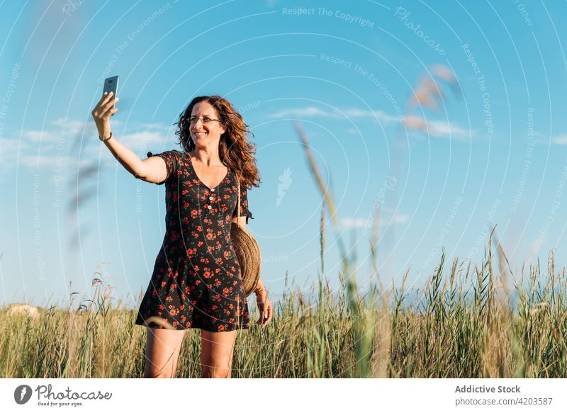 Smiling woman taking selfie on smartphone in nature summer field smile dress sunset self portrait female moment memory take photo enjoy gadget device delight