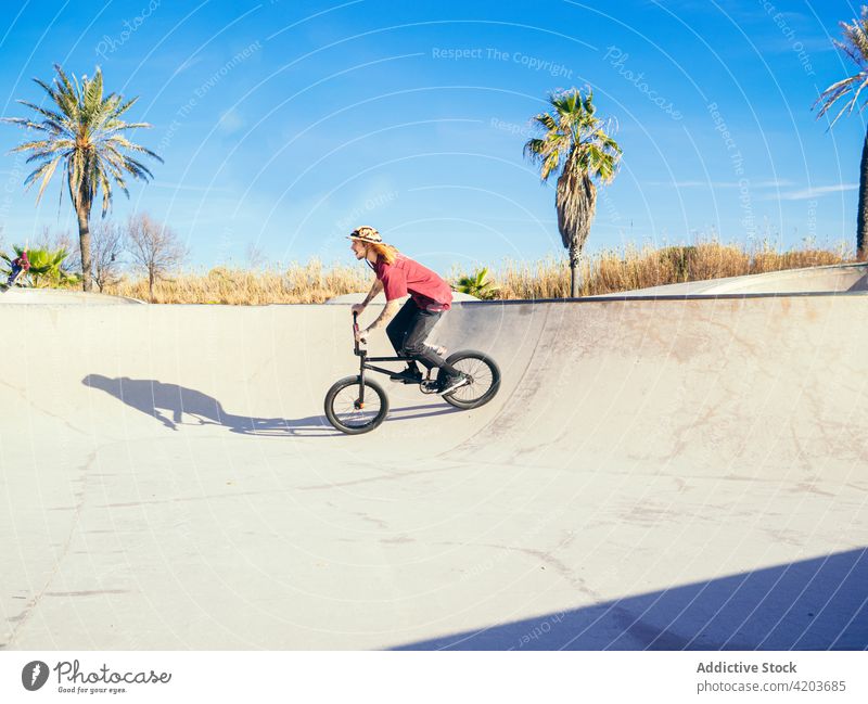 Sportsman riding trial bike in skate park biker ride sport training workout active bmx blue sky cloudy tropical exotic palm tree shadow bicyclist ramp hipster