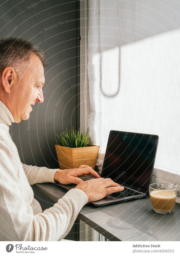 Cheerful mature man siting on chair at counter with laptop in kitchen morning smile home breakfast coffee male work cup netbook remote cheerful happy middle age