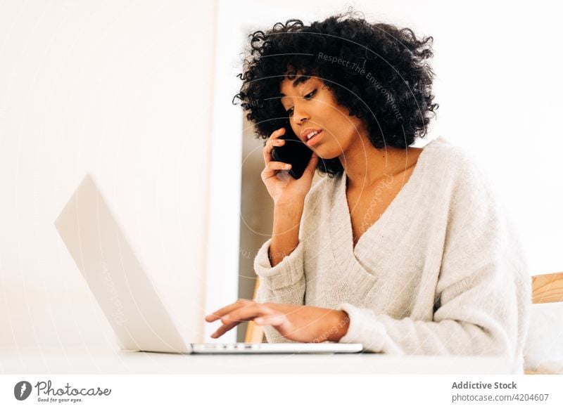 Black woman talking on smartphone and working on laptop at home multitask freelance remote project busy female ethnic black african american table typing