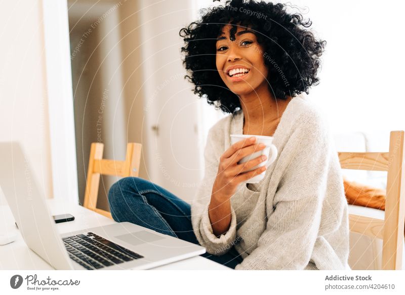 Content black woman sitting at table with laptop and cup of drink freelance cheerful home office beverage entrepreneur project female ethnic african american
