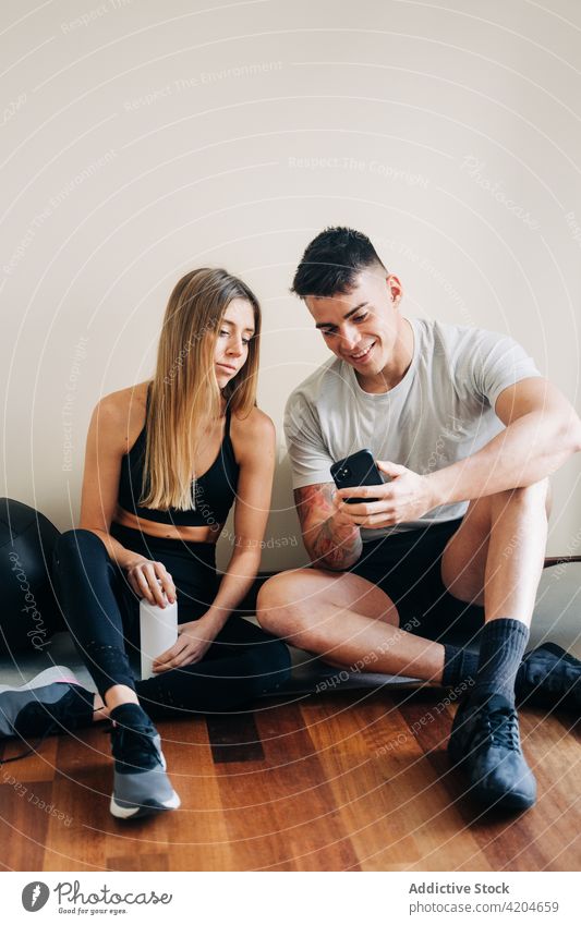 Cheerful couple browsing cellphone after training smartphone bottle water rest wall hydrate internet workout sport fitness practice woman activewear abs athlete