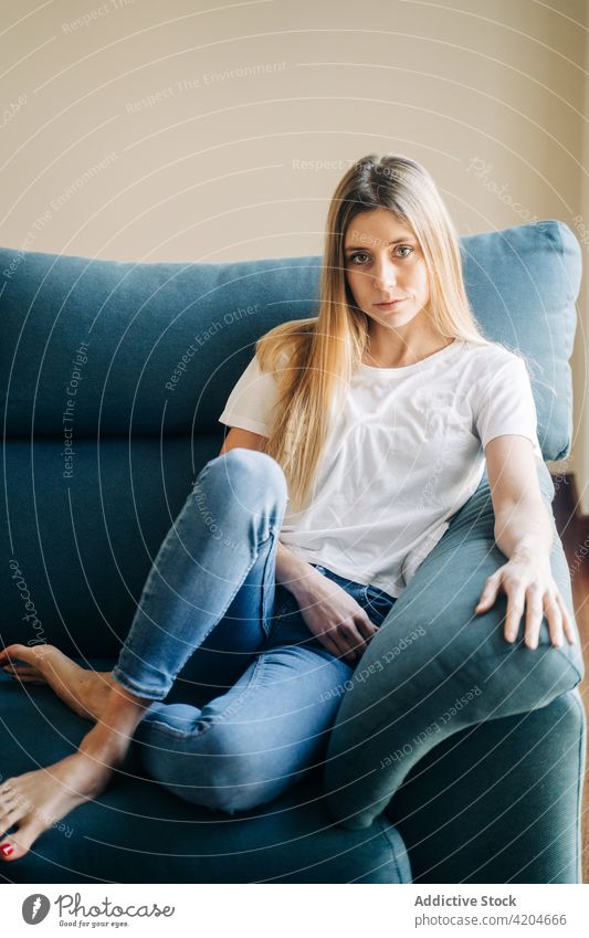 Serious woman sitting on couch sofa legs crossed barefoot living room day off domestic home cushion female jeans lady lifestyle calm serious flat pensive