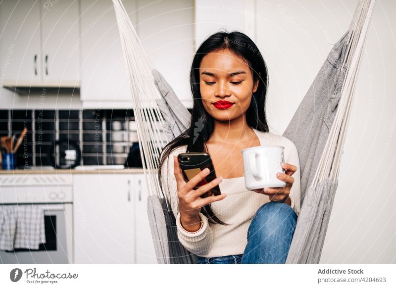 Hispanic lady text messaging via smartphone while sitting in hammock woman beverage using social media online browsing device female hispanic mobile young