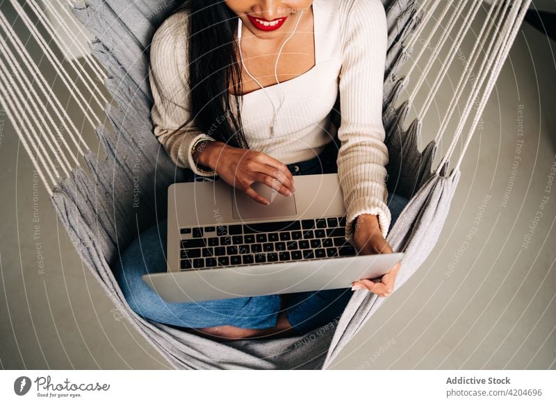 Cheerful young woman working on laptop while sitting in hammock freelance smile browsing gadget social media earphones using comfort netbook female surfing