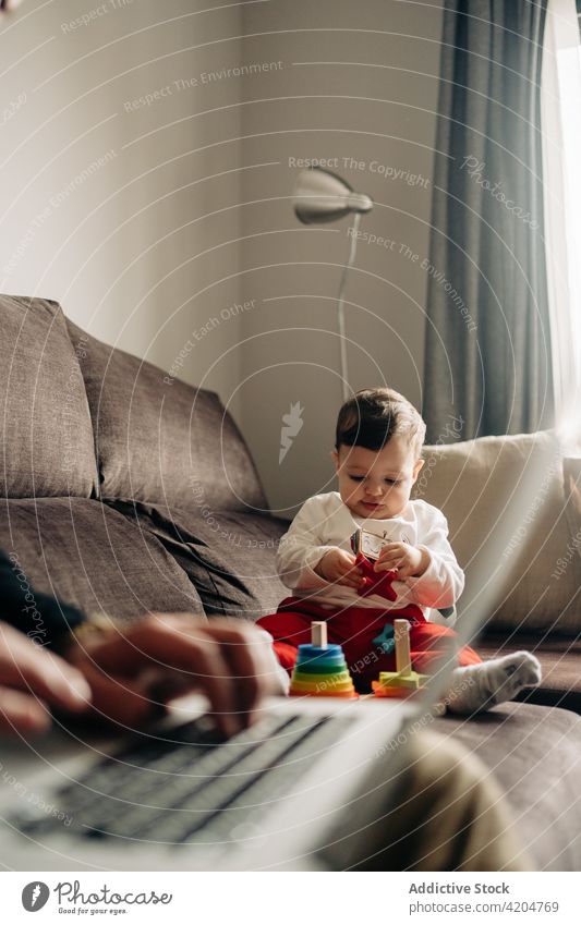 Curious baby playing with toys on couch near anonymous father using laptop sofa cone tower typing together playful curious colorful wooden toddler boy child