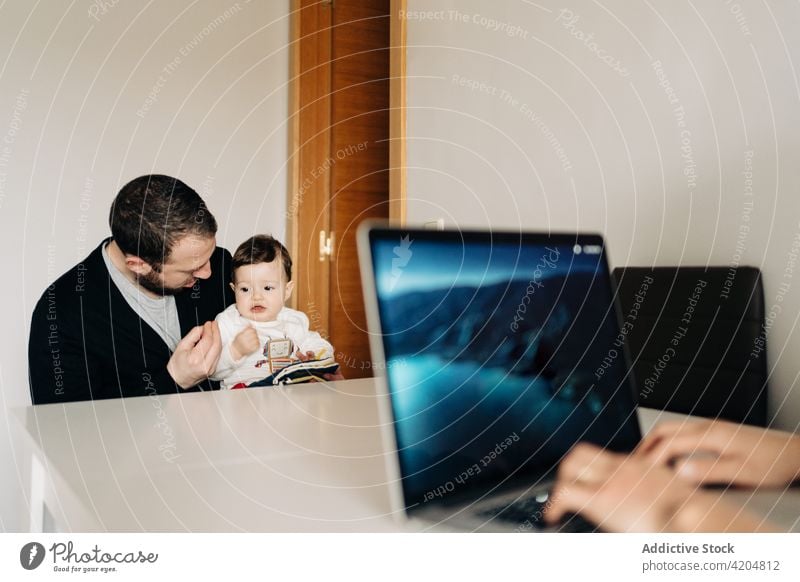 Father and baby reading book near mother working on laptop family freelance browsing childcare parenthood development together young father cute kid love