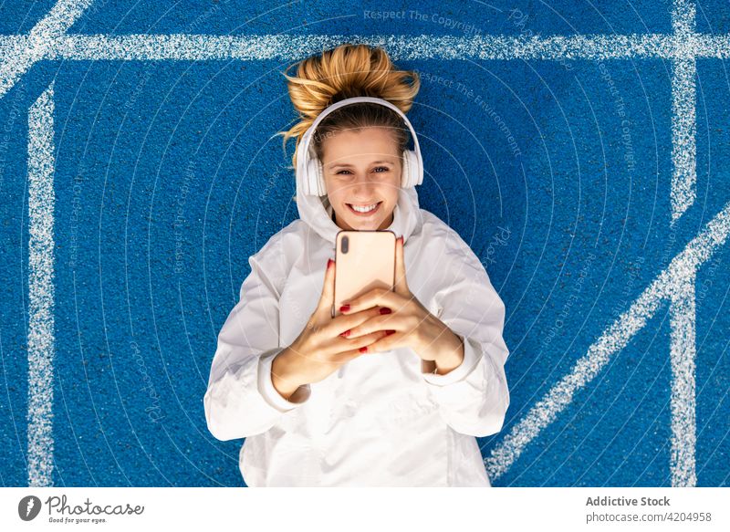 Content sportswoman taking selfie on racetrack at stadium athlete smartphone self portrait track and field headphones music female smile lying blue color