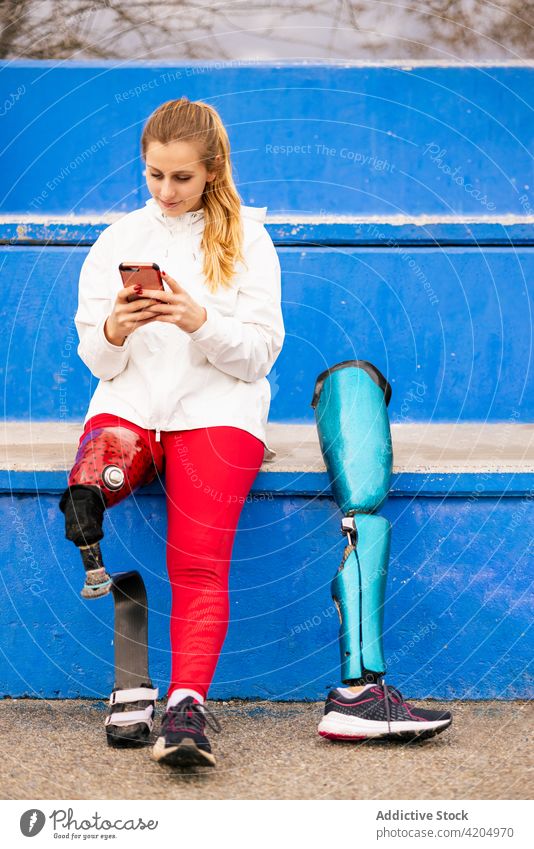 Sportswoman with bionic prosthesis using smartphone at stadium sportswoman browsing leg athlete training break female paralympic artificial limb professional