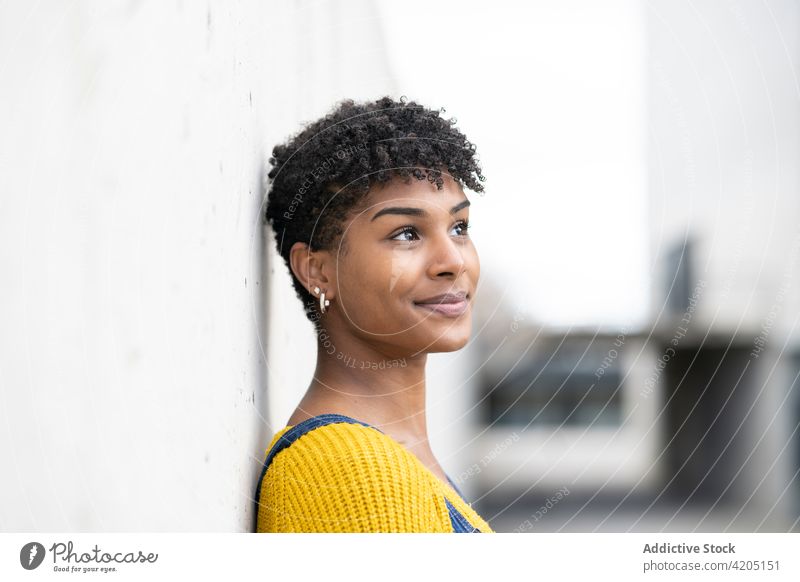 Delighted ethnic woman on street and looking away smile afro hairstyle city urban overall outfit cheerful female black african american wall lean positive