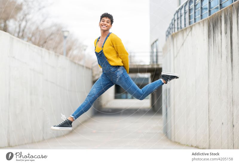 Carefree black woman jumping in city above street carefree moment cheerful freedom leap energy female ethnic african american overall denim sneakers cool smile