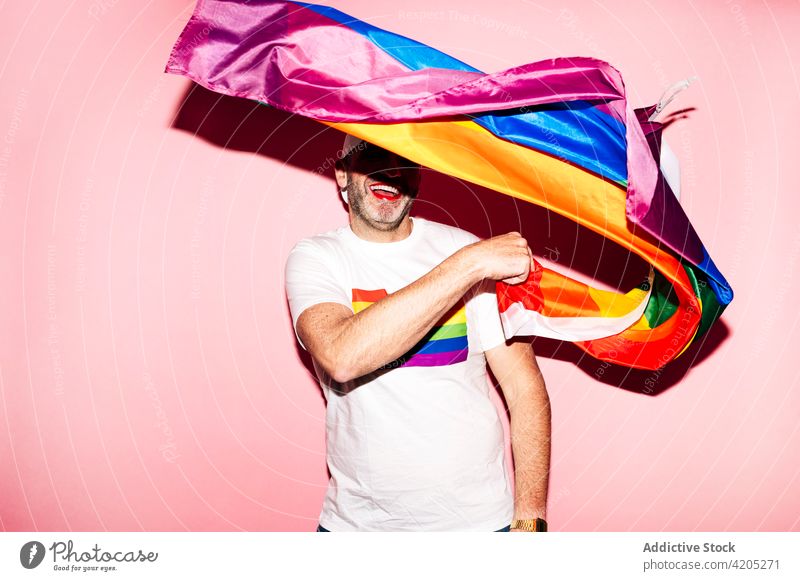 Happy man with makeup waving LGBT flag in pink studio homosexual lgbt cheerful happy gay smile concept pride rainbow equal male unshaven red lips respect