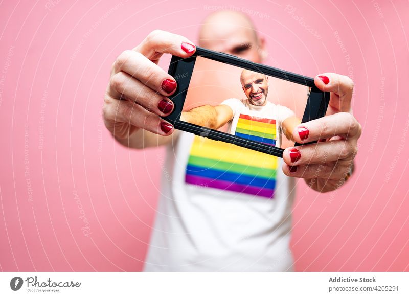 Cheerful male in shirt with LGBT flag taking selfie on smartphone man lgbt gay eccentric smile queer concept equal homosexual happy bald beard red lips manicure