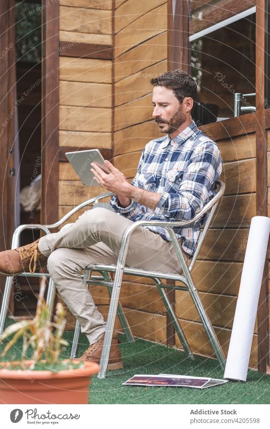 Male architect using tablet on terrace of house man project work browsing blueprint draft male paper roll whatman sketch wooden sit chair communicate gadget