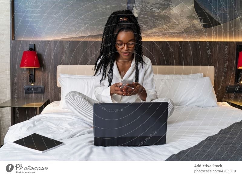 Self employed young black lady using laptop and smartphone on bed while working distantly woman freelance busy remote internet online multitask job female