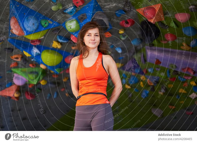 Sportswoman standing near climbing wall in gym sportswoman hands behind back mountaineer climber smile confident fit fitness healthy athlete vitality energy