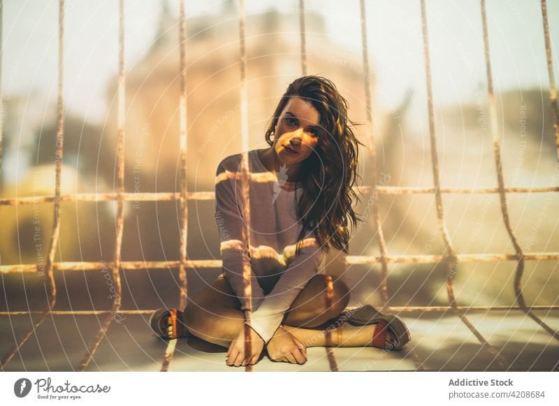 Young woman under projection of cage concept captive summer grate bar outfit legs crossed female trap lonely alone solitude long hair casual enclosure