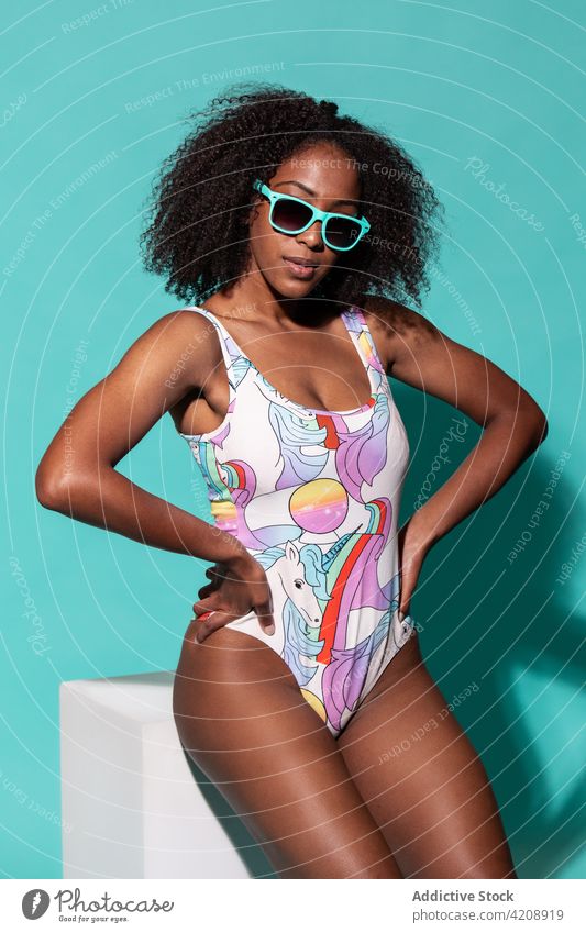 Gorgeous black woman in swimsuit sitting in bright studio swimwear style hand on waist posture cheerful outfit sunglasses cool charismatic carefree colorful