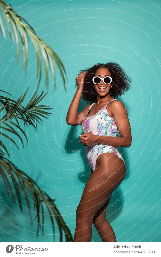 Cheerful black woman in swimsuit standing on blue background swimwear style happy touch hair cheerful sunglasses cool charismatic joy outfit carefree colorful