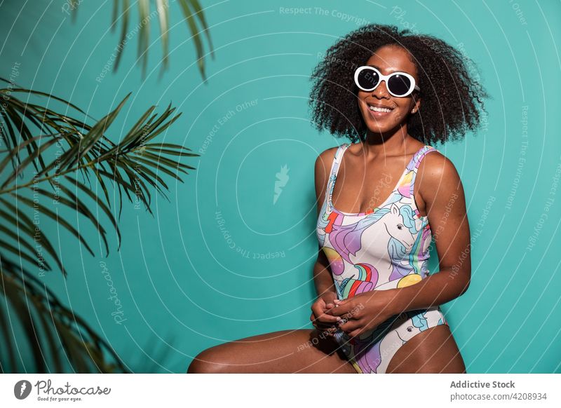 Cheerful black woman in swimsuit sitting on blue background swimwear style happy cheerful sunglasses cool charismatic joy outfit carefree colorful satisfied