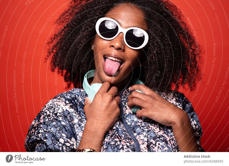 Stylish black woman with headphones on red background cool tongue out music arms raised outfit style grimace make face happy entertain song meloman melody sound