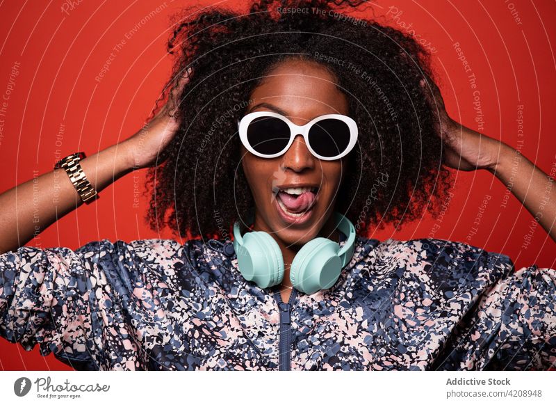 Stylish black woman with headphones touching hair on red background cool tongue out music arms raised touch hair outfit style grimace make face happy entertain