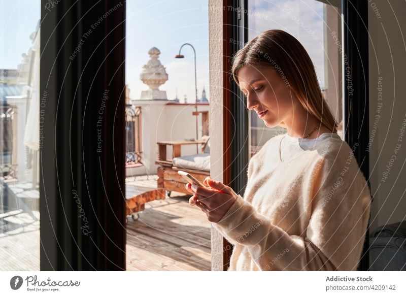Pensive young woman using smartphone near terrace at home message window communicate chat internet online connection social media mobile female sweater style