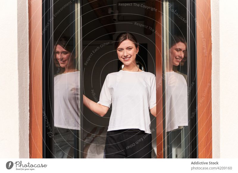 Joyful young woman smiling while standing near glass doors at home smile positive balcony content cheerful reflection happy glad joy female casual delight rest
