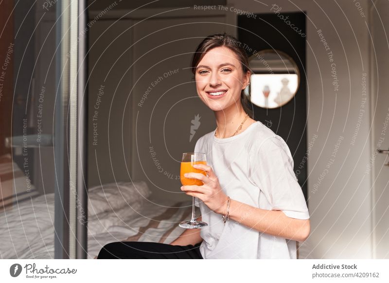Happy young woman drinking juice on bed at home content morning chill fresh healthy beverage comfort cozy smile female casual window balcony reflection optimist