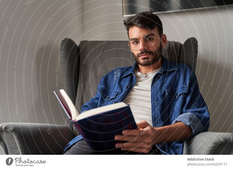 Serious young ethnic man sitting in armchair and reading novel book home relax knowledge literature hobby concentrate male casual hispanic interesting daylight