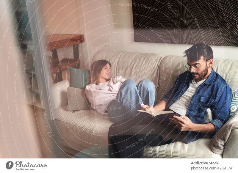 Serious Hispanic man reading book on sofa near sleeping girlfriend couple rest literature relationship together weekend eyes closed lying knowledge smart