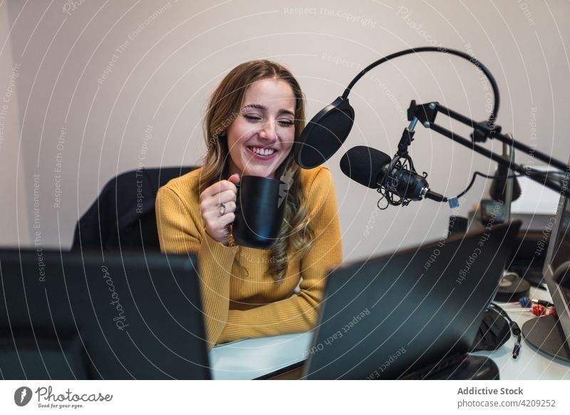 Cheerful woman with coffee talking to microphone at broadcast studio work radio record laptop cup mug hot drink equipment speak device special modern netbook