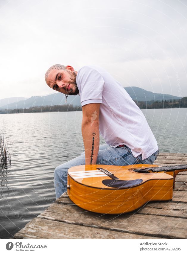 Modern man with guitar on pier near lake river nature musician mountain wooden cloudy gray sky guitarist daytime peaceful water summer alone plant guy shore