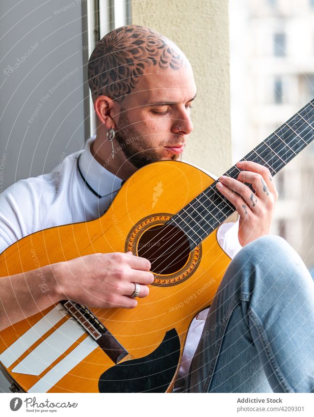 Musician sitting near window and playing guitar in daytime man musician tattoo melody hobby mandala pensive perform male guitarist focus bald windowsill