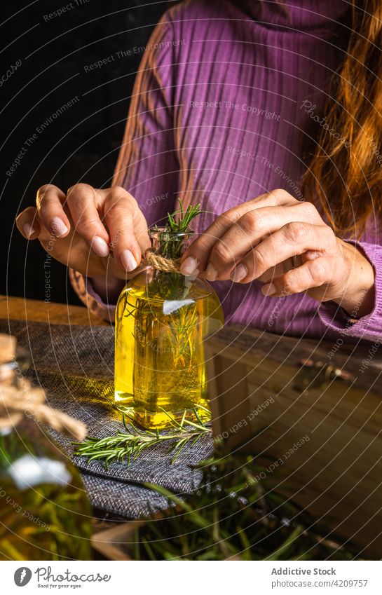 Unrecognizable female demonstrating bottle with oil and rosemary woman herb demonstrate cloth table glass show delicate essential light purple casual creative