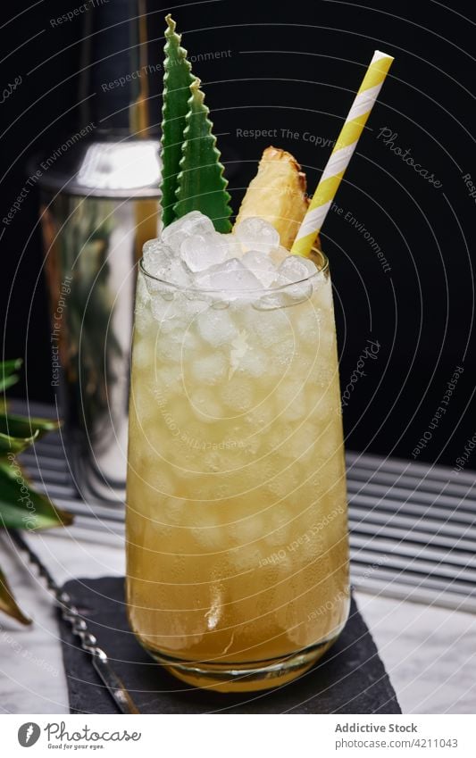 Glass with pineapple cocktail near shaker ice slate cube straw counter spoon leaf glassware creative fresh paper coaster exotic ingredient cold fruit yummy