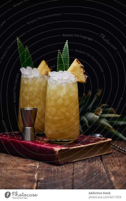 Glasses with cocktails with pineapple pieces on book glass ice refreshment table leaf drink spoon beverage fruit alcohol tasty cold yummy portion ingredient