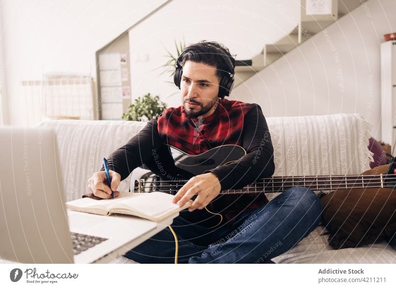 Bassist with guitar writing in notepad against laptop in house bassist write compose create headphones listen man using gadget device take note diary sofa