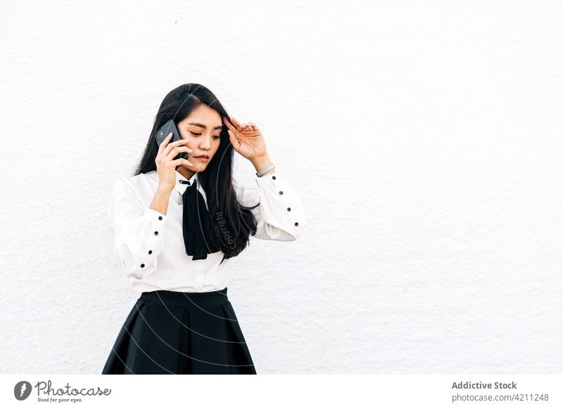 Asian woman speaking on smartphone against white wall student phone call using teen talk internet communication elegant thoughtful positive connection formal