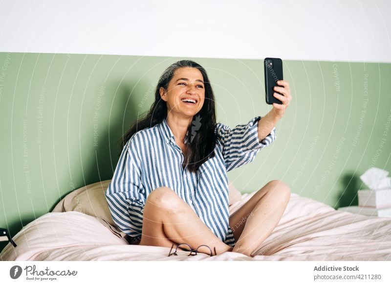 Content mature Hispanic woman taking selfie on bed smile smartphone confident cheerful relax bedroom social media delight moment female middle age ethnic