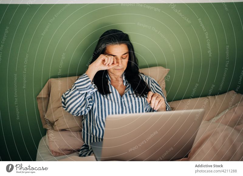 Tired ethnic woman rubbing eyes during online work on laptop on bed exhausted remote freelance rub eye tired overwork job female middle age hispanic home