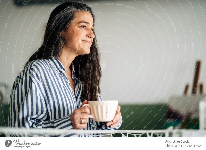 Positive mature woman with coffee in room cup domestic drink leisure recreation painting middle age easel drawing female hot drink beverage smile home enjoy