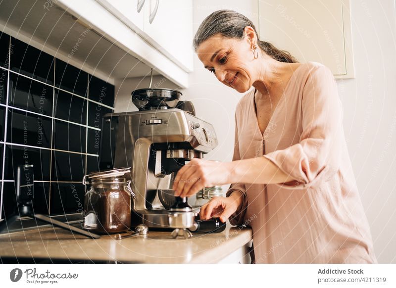 Focused mature woman preparing beverage with coffee machine coffeemaker prepare smartphone cup kitchen equipment kitchenware domestic drink cupboard middle age