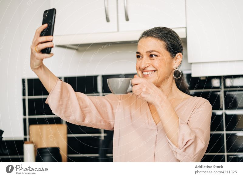 Cheerful mature woman with coffee taking selfie in kitchen self portrait smartphone cup photography drink cupboard domestic cellphone middle age female smile