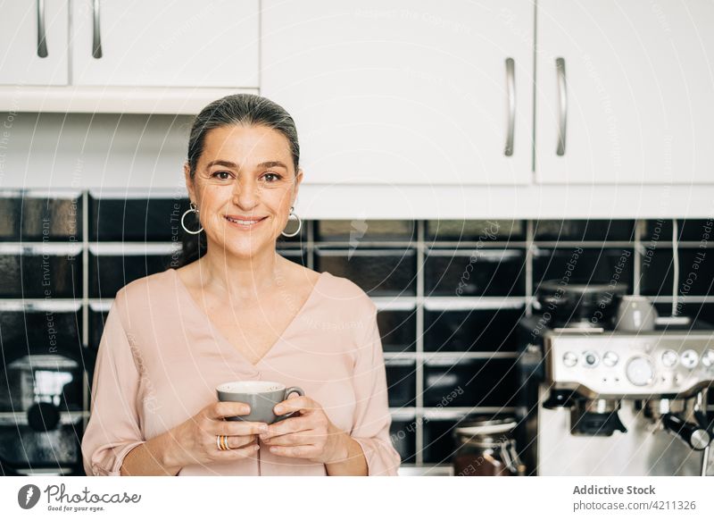 Happy mature woman with cup of coffee in kitchen coffeemaker coffee machine equipment drink domestic kitchenware cupboard middle age female smile home enjoy mug