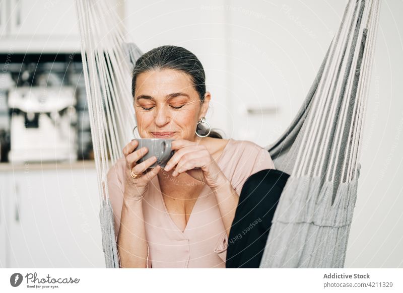 Cheerful mature woman drinking coffee while sitting on hammock kitchen cup room domestic leisure recreation middle age female eyes closed hot drink beverage