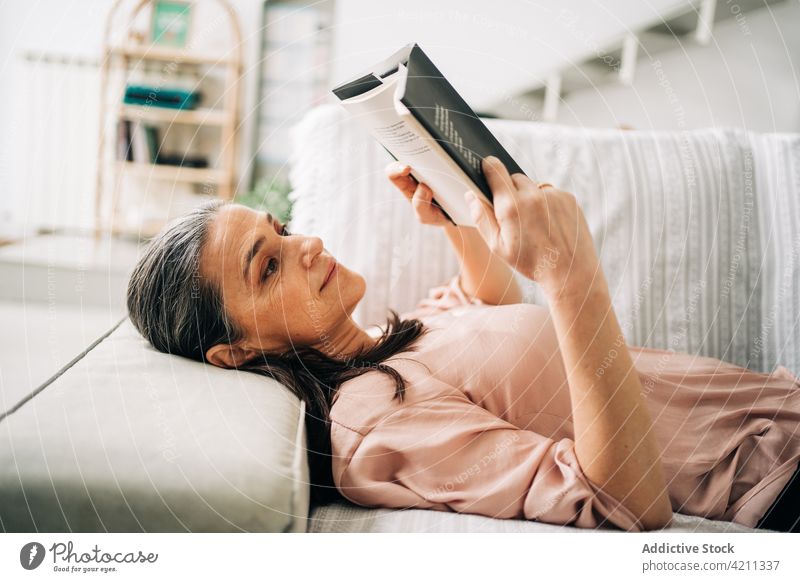 Concentrated woman reading book on couch sofa apartment domestic hobby bookworm living room lying down female home casual concentrate comfort flat focus plant