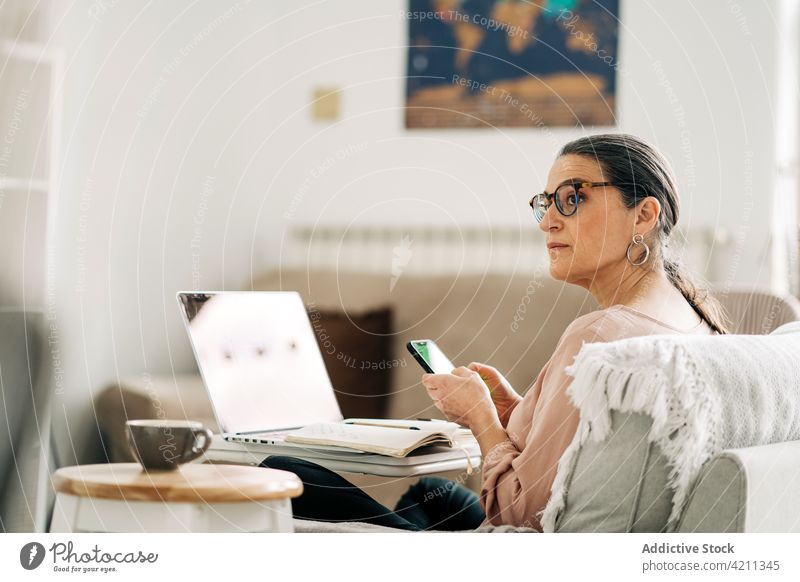 Serious mature woman on mobile phone and working on computer smartphone communicate laptop internet browsing middle age message online sofa room female